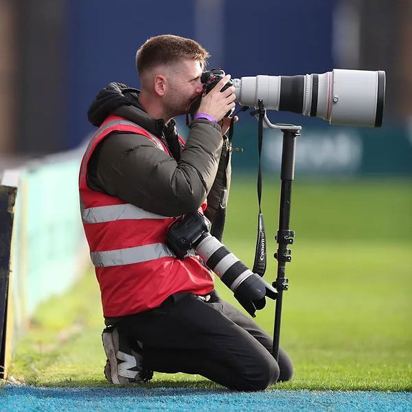 A sports photographer kneeling on the ground, capturing images with a large camera lens.