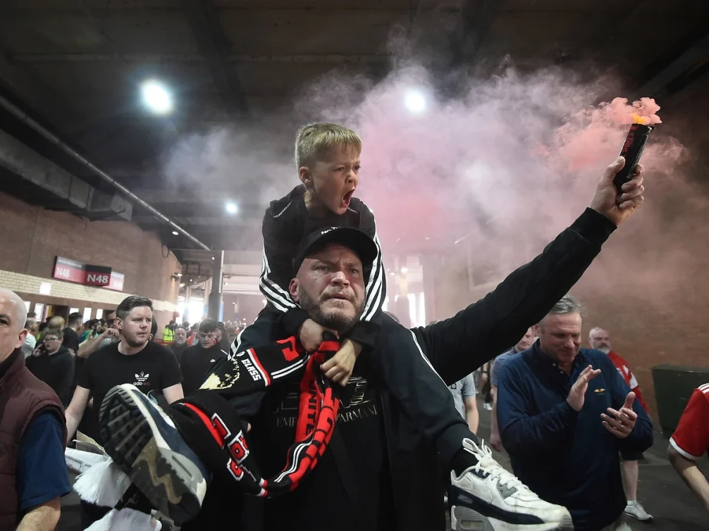 A father carrying his son on his shoulders, both celebrating passionately during a football match.